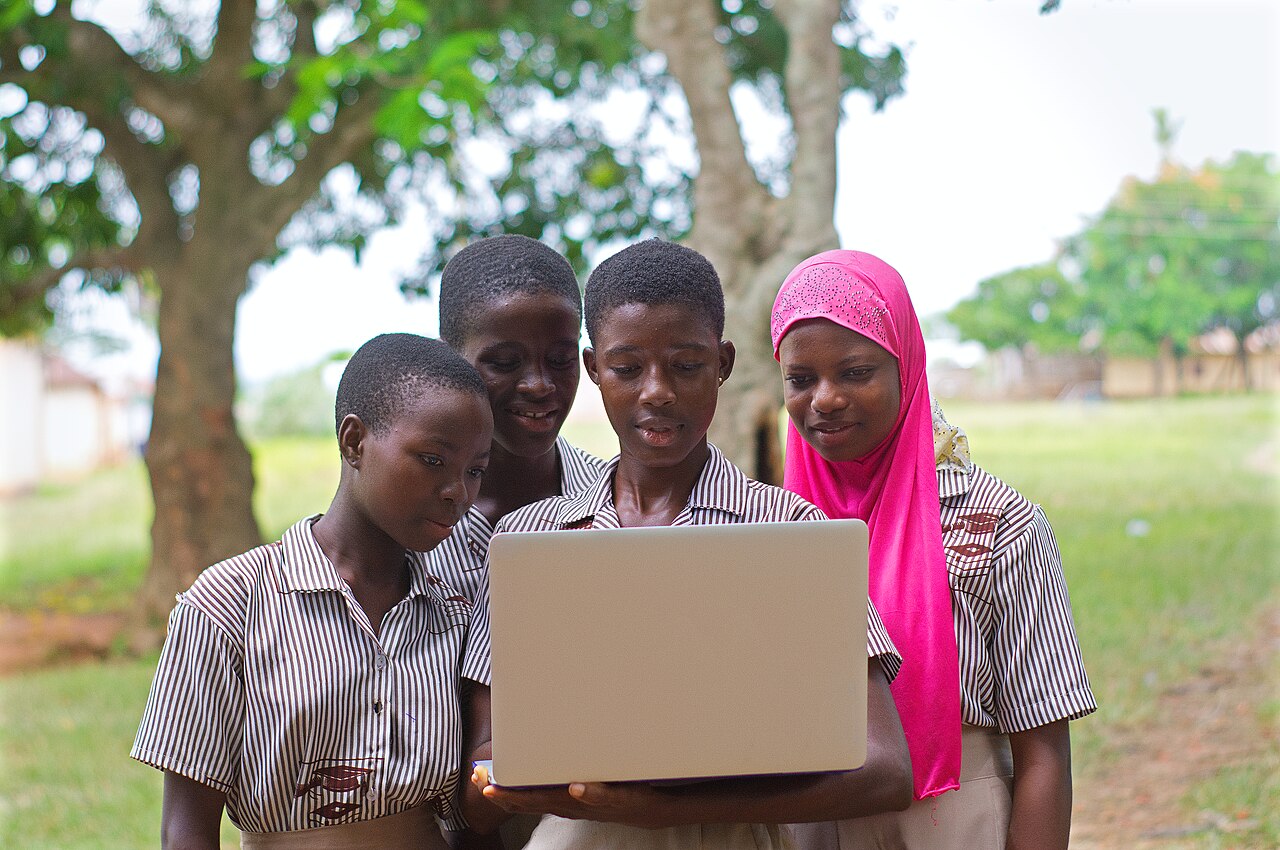 Four junior-high students gather around a laptop outdoors and look closely at its screen.